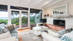 A spacious living room with a fireplace, a television, and a large window overlooking greenery. - The Grove (Lorne)