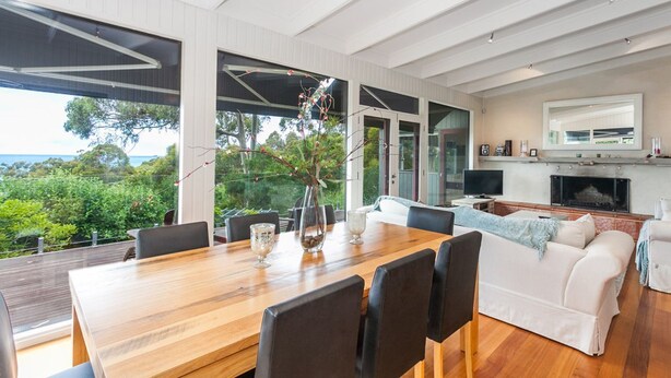 A modern living room with a wooden dining table, comfortable seating, a fireplace, and a large window offering a view of the outdoors. - The Grove (Lorne)