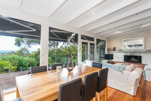 A modern living room with a wooden dining table, comfortable seating, a fireplace, and a large window offering a view of the outdoors.