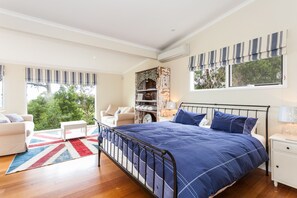A bedroom with a large bed, a white nightstand, a white dresser, a window with striped valance, and a view of greenery outside.