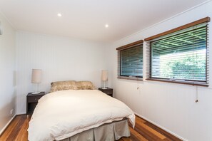 A bedroom with a large bed, two bedside tables, a window with blinds, and a radiator. - The Grove (Lorne)