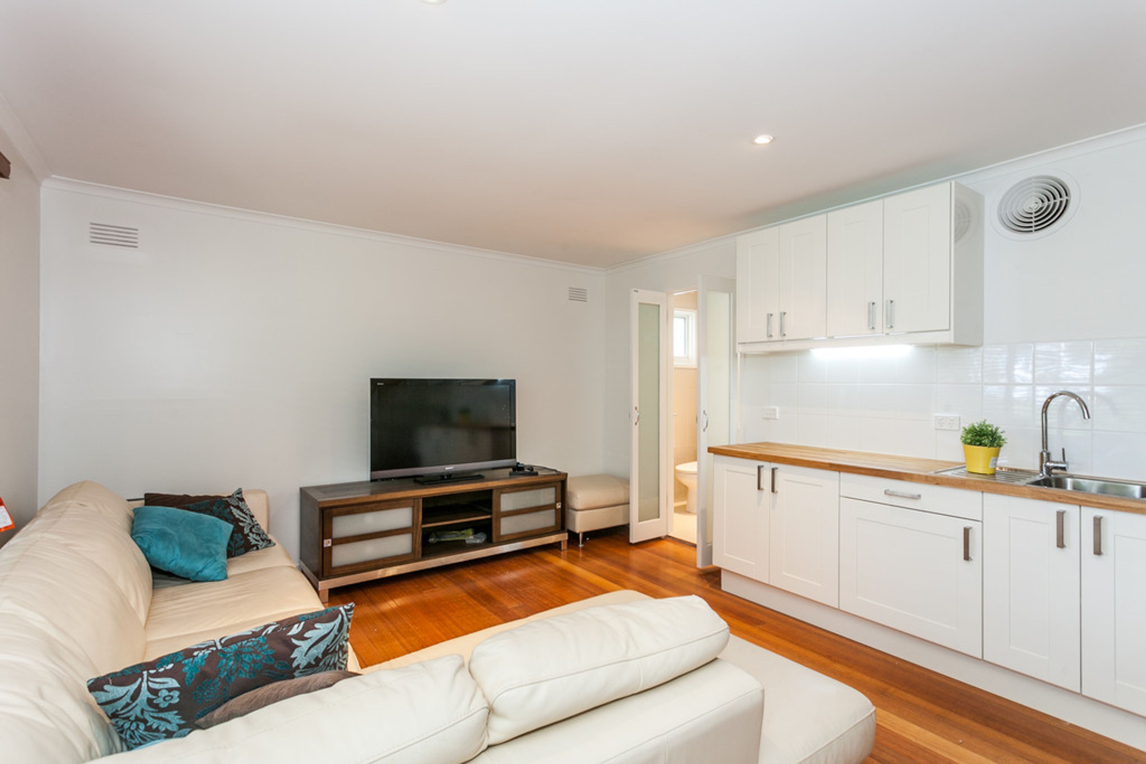 A modern living room with a sofa, a TV, and a kitchen with white cabinets and a wooden countertop.