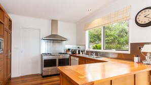 A modern kitchen with stainless steel appliances, wooden cabinetry, a large window with a view of greenery, and a clock on the wall. - The Grove (Lorne)