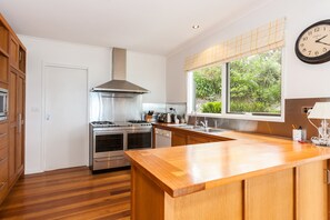 A modern kitchen with stainless steel appliances, wooden cabinetry, a large window with a view of greenery, and a clock on the wall. - The Grove (Lorne)