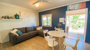 A living room with a dark grey sofa, white chairs, a round table, and a ceiling fan. - The Batch - Burrum Heads - Walk To Beach - 2BR - Pets Welcome (BURRUM HEADS)