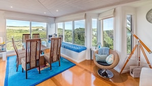 A spacious room with large windows offering a view of the countryside. There is a dining area with a table and chairs, a seating bench with cushions, and a wicker chair. - Mills Court, Quirky seaside house at Smiths Beach, bring the surf boards! (Smiths Beach)