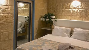 Un dormitorio con pared de piedra, cama con colcha estampada, mesita de noche con planta y espejo en la pared.