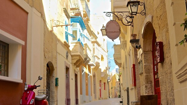 Una estrecha calle adoquinada flanqueada por edificios históricos, con una scooter roja estacionada al lado y un letrero colgante que indica un restaurante.
