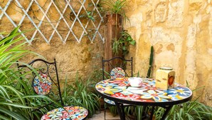 Un pequeño patio al aire libre con una mesa redonda y dos sillas, rodeado de plantas en macetas y un muro con celosía.