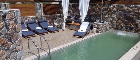 En pool med oändlig horisont, omgiven av stenmurar, med solstolar och en pergola med vita draperier.
