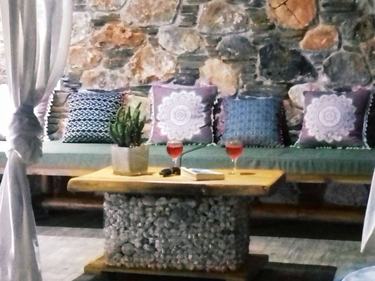 Un mur de pierres, avec un canapé et des coussins, une table sur laquelle se trouvent une plante et deux verres contenant un liquide rouge.
