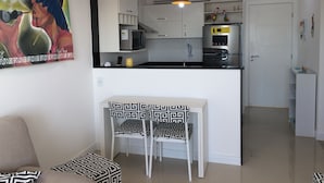 Una cocina moderna con armarios blancos, un microondas y un refrigerador. Hay un comedor con una mesa y sillas, y un cuadro en la pared.