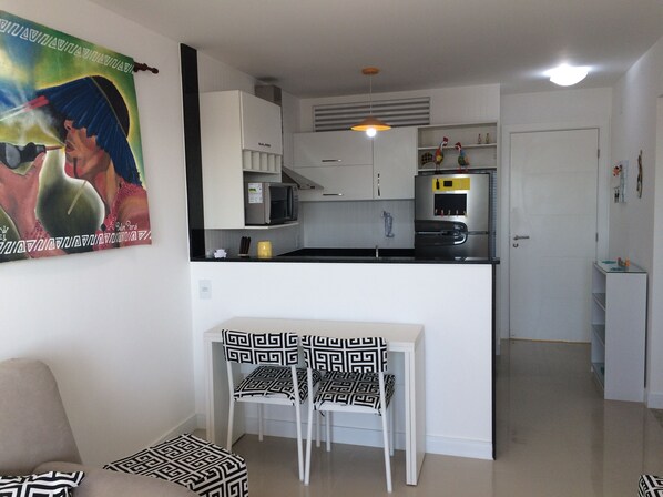A modern kitchen with white cabinets, a microwave, and a refrigerator. There is a dining area with a table and chairs, and a painting on the wall.