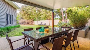 An outdoor patio with a glass-top table and wicker chairs, surrounded by a garden and a house.