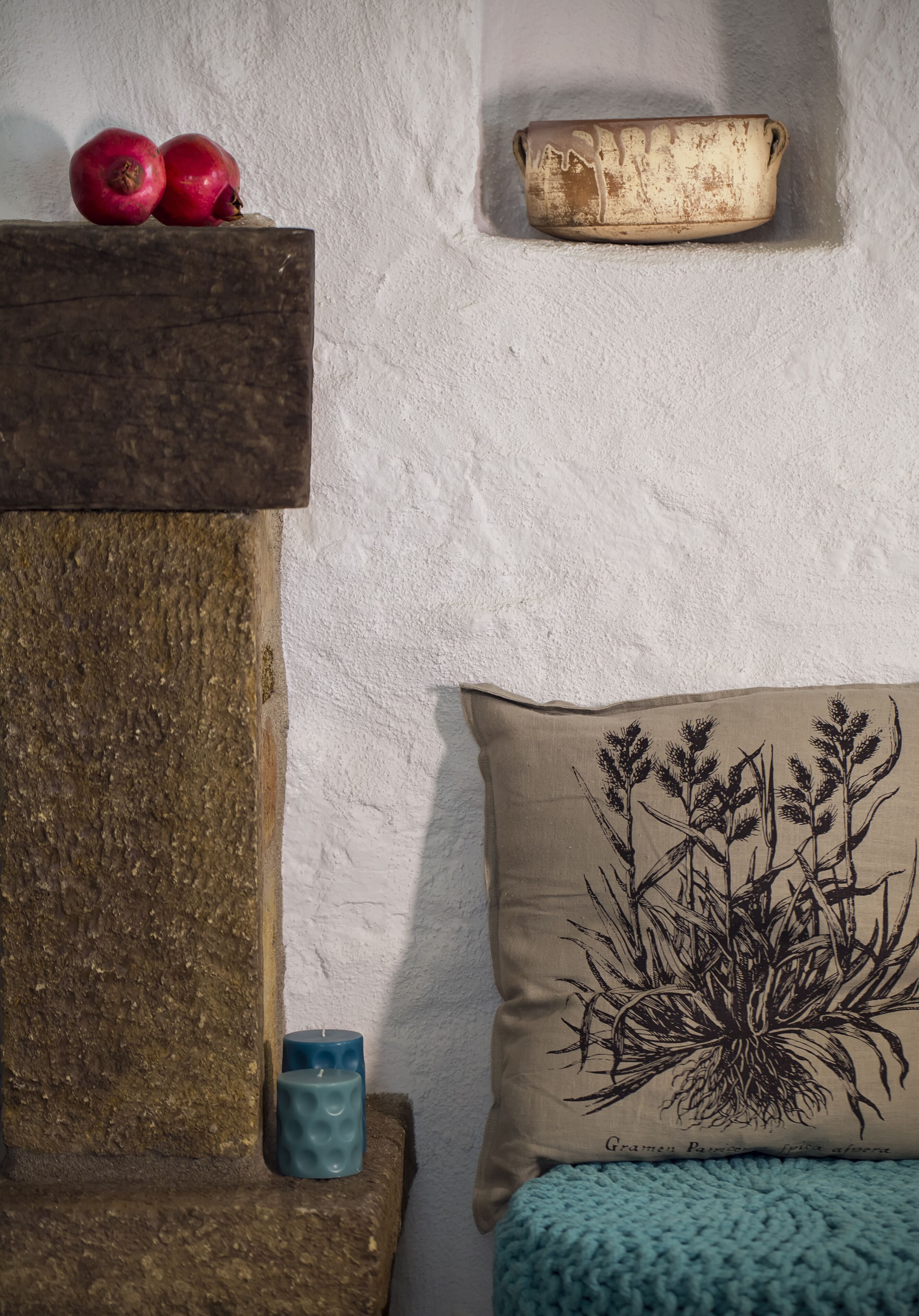 A rustic room with a stone shelf holding two pomegranates, a textured wall, a woven basket, and a cushion with a plant design.