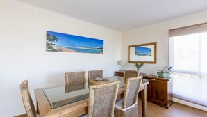 Een eethoek met rieten stoelen, een tafel met glazen blad en een schilderij van een strand.