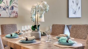 Una mesa de comedor lista, con platos, vasos y un jarrón de flores blancas.