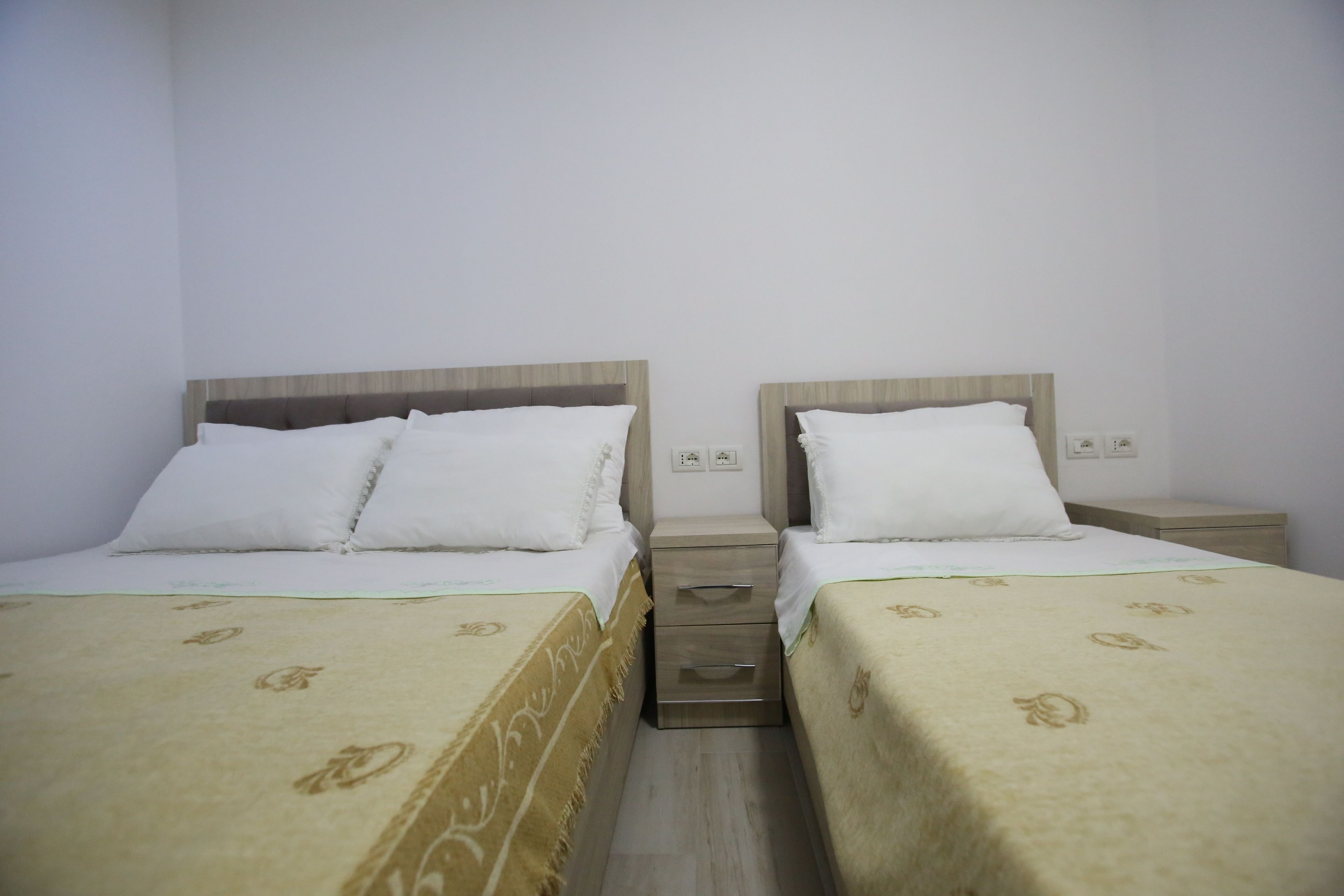 A hotel room with two single beds, each with a wooden headboard, white bedding, and a beige patterned bedspread. There is a small bedside table with a lamp between the beds.