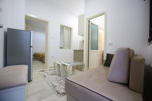A compact living space with a sofa, a small table, a refrigerator, and a glass door leading to another room. - Idrizi Apartment (Berat)