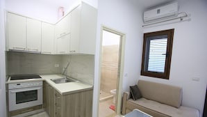 A compact kitchen with a built-in oven, a sink, and wooden cabinets. There is a beige sofa and a small table in the living area, and a window with blinds. - Idrizi Apartment (Berat)