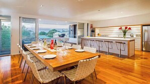 A modern dining room with a wooden table and white chairs, a kitchen with a central island, and large windows offering a view.