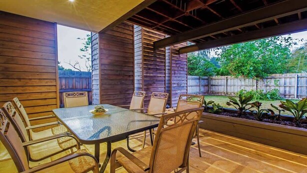 An outdoor patio with a glass-top table and wicker chairs, surrounded by wooden walls and a view of a backyard.