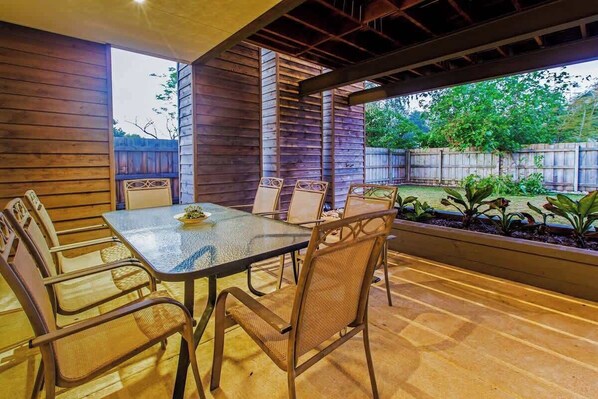 Une terrasse extérieure dotée d’une table à plateau de verre et de chaises en osier, entourée de murs en bois et offrant une vue sur un jardin.