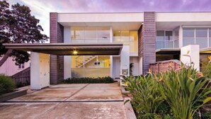 A modern two-story house with a covered driveway, a staircase leading to the upper level, and a well-maintained garden with tropical plants.