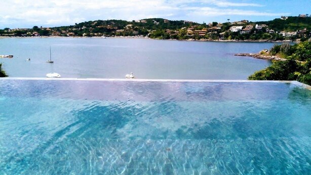 Una piscina infinita de color azul claro con vistas a un mar tranquilo y un pueblo en la colina al fondo.