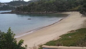 Vista costera con una playa arenosa, una escalera que baja a la playa y una colina con edificios al fondo.