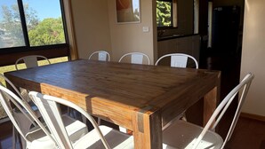 A dining table with chairs, a kitchen area, and a window with a view of trees. - Shelby s Lodge- lovely light filled home (Venus Bay)
