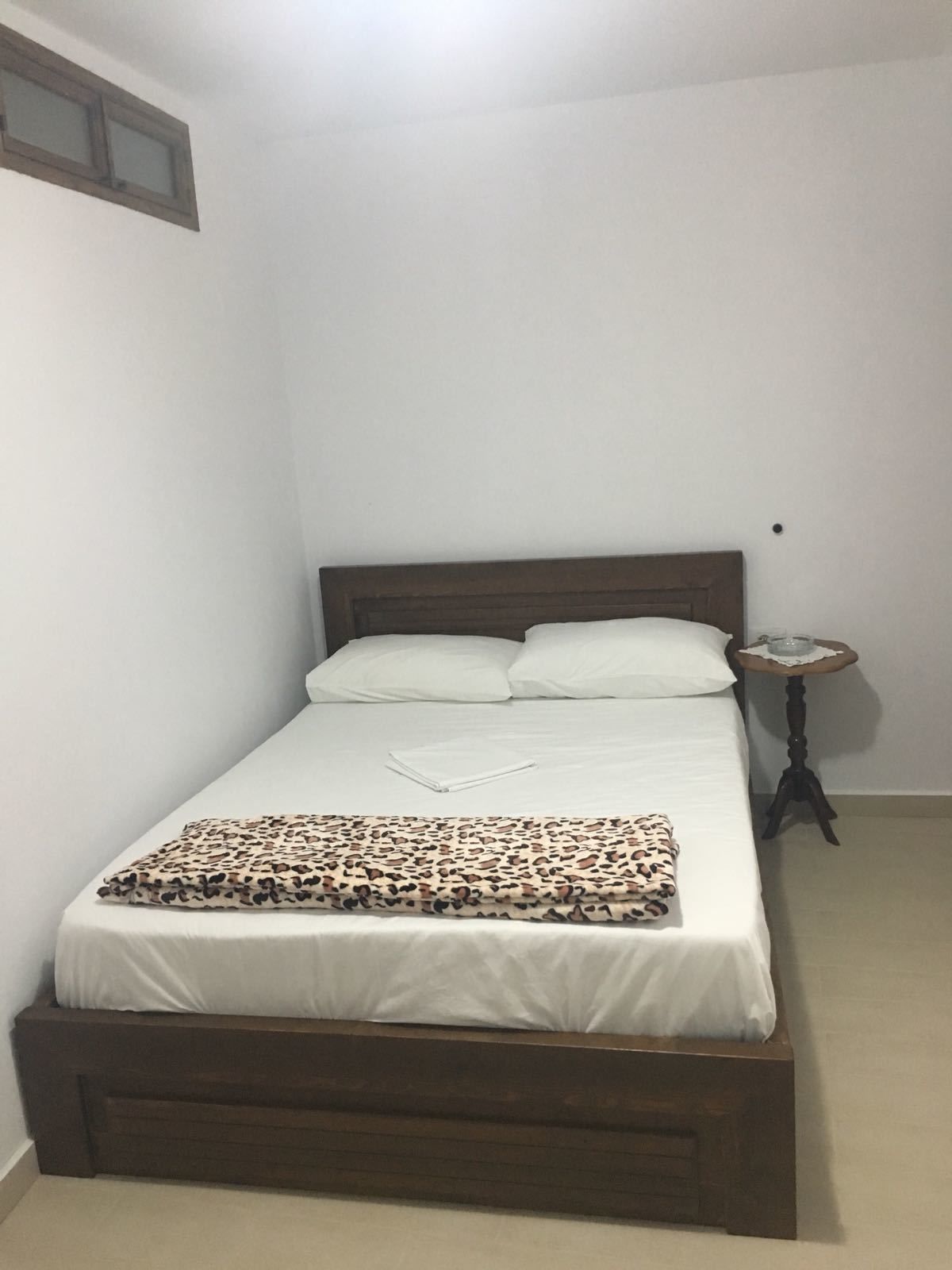 A single bed with a wooden headboard, white bedding, and a leopard print blanket. There is a small bedside table with a glass and a book.