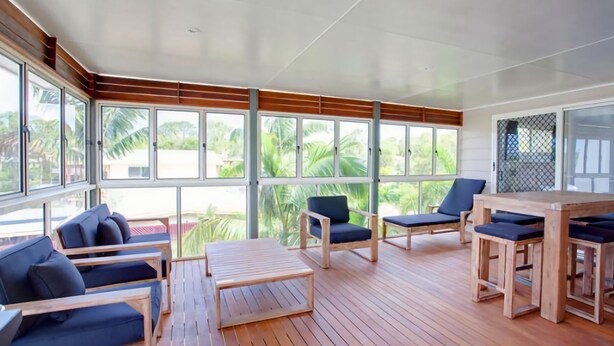 A spacious outdoor area with wooden flooring, multiple seating arrangements including sofas and chairs, and large windows offering views of greenery. - 29 Cypress Avenue - Rainbow Beach. Close to the beach with a pool (RAINBOW BEACH)
