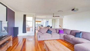 A modern living room with a sectional sofa, wooden flooring, a flat-screen TV, and a dining area in the background. - 29 Cypress Avenue - Rainbow Beach. Close to the beach with a pool (RAINBOW BEACH)
