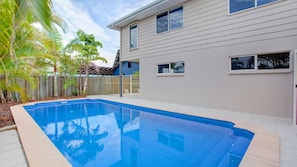 A residential swimming pool with a wooden deck, surrounded by a fence and palm trees.