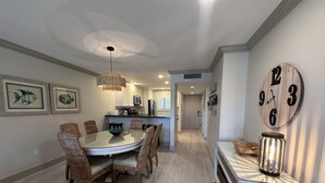 A dining area with a round table and wicker chairs, a kitchen in the background, and a wall clock. - Updated Gulf View Condo in Beautiful Siesta Dunes, 5-205 (Siesta Key)