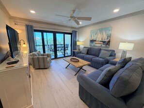 A living room with a sofa, armchair, and a coffee table. There is a ceiling fan, a painting on the wall, and a view through the glass doors. - Updated Gulf View Condo in Beautiful Siesta Dunes, 5-205 (Siesta Key)