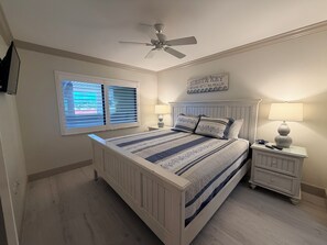 Ein Schlafzimmer mit einem Bett, zwei Nachttischen, einem Deckenventilator, einem Fenster mit Jalousien und einem Wandschild.