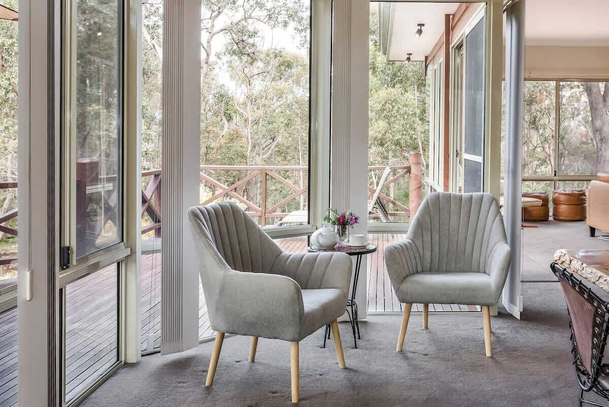 A room with two grey upholstered chairs, a small table with a vase, and large windows offering a view of trees.