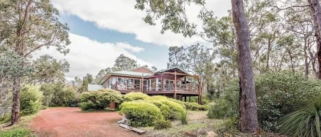 A two-story wooden house with a deck, surrounded by dense greenery and trees.