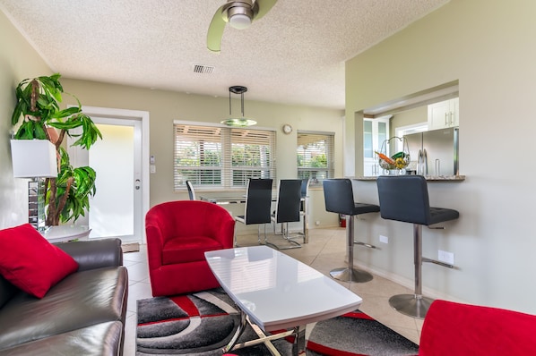 A modern living room with a red sofa, a white coffee table, and a red armchair. There is a large potted plant, a dining area with a bar stool, and a kitchen area visible through the window.