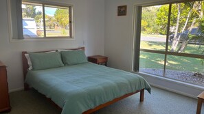A bedroom with a bed, a nightstand, and a window with a view of a parking lot and trees. - Baker - Rainbow Beach. Walk to Everything! Including the beach and main street. (Rainbow Beach)