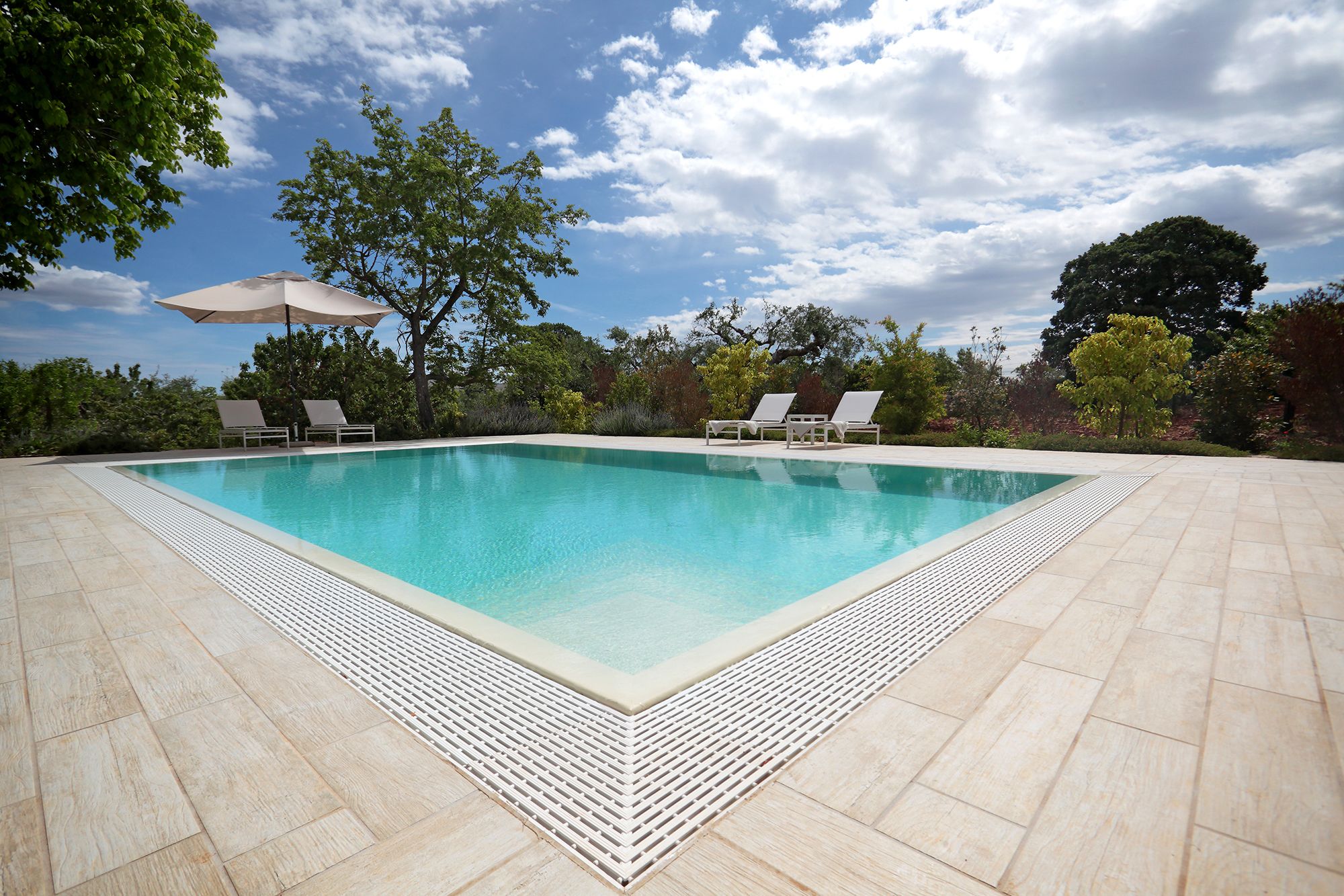 Una piscina rectangular con un borde empedrado, rodeada de árboles y un cielo despejado.