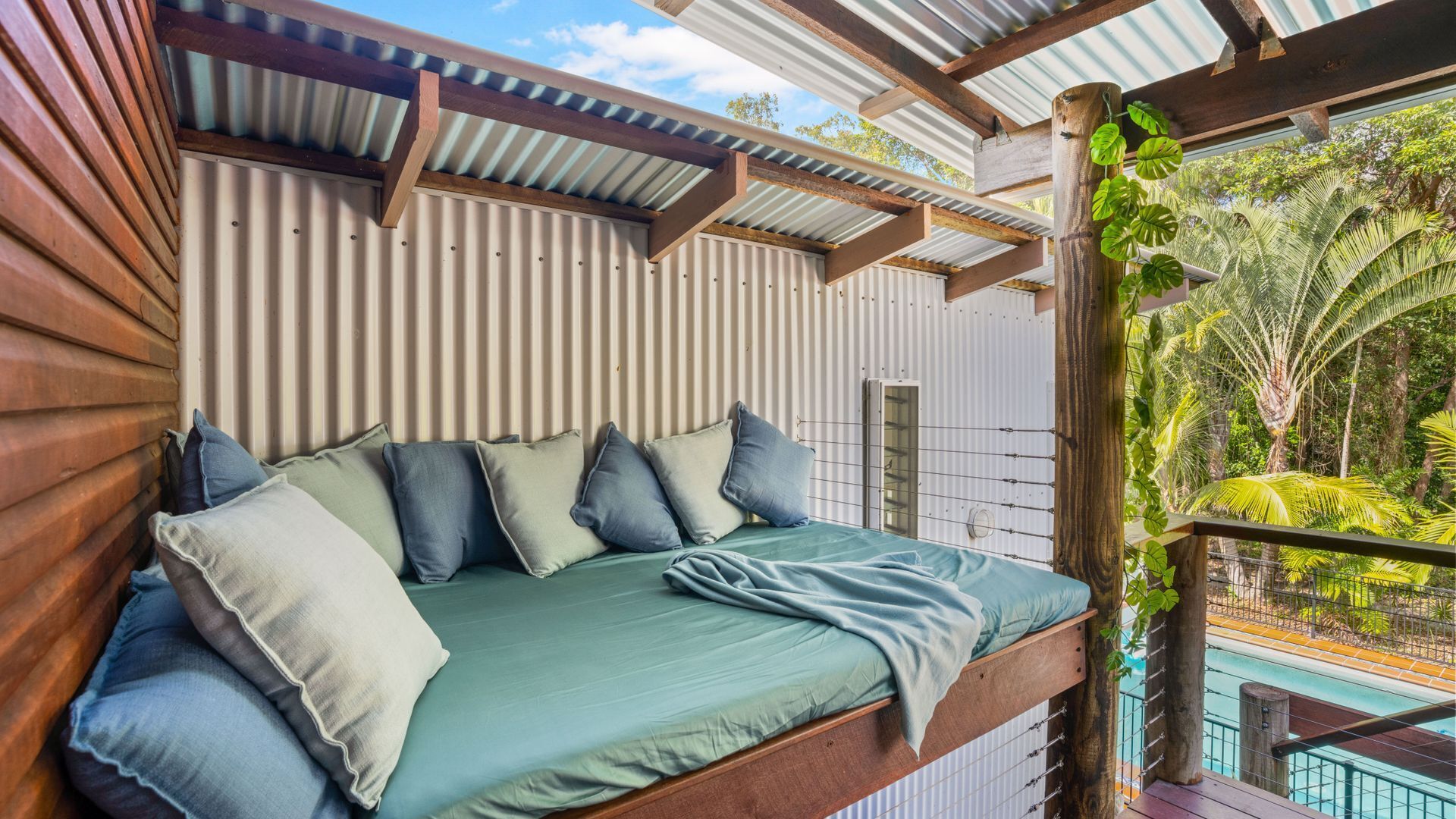 A bed with a green mattress and multiple pillows, a wooden headboard, and a view of a pool and greenery.