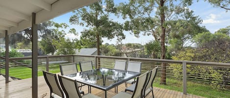 A covered outdoor deck with a glass-top table and chairs, surrounded by a wooden deck and a view of trees and a distant building.