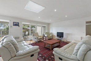 A spacious living room with a large sofa, a smaller sofa, a coffee table, and a television. There is a sliding glass door leading to an outdoor area with chairs. - Beach Side (Aireys Inlet)