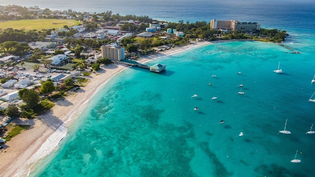 Luchtbeeld van een kustresort met een strand, helder turquoise water en verschillende boten.
