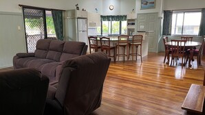 Een woonkamer met een bruine bank, een eethoek met houten stoelen en een tafel, en een keuken met witte kastjes en een raam.