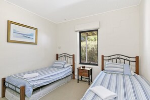 A bedroom with two single beds, a small bedside table, a window with a view of greenery, and a framed picture on the wall. - Fernview (Lorne)