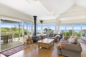 A spacious living room with a large fireplace, comfortable seating, and a view of the ocean. - Fernview (Lorne)